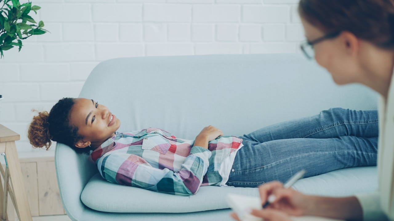 our-experience A woman in therapy, smiling while lying on a couch, with a counselor taking notes.